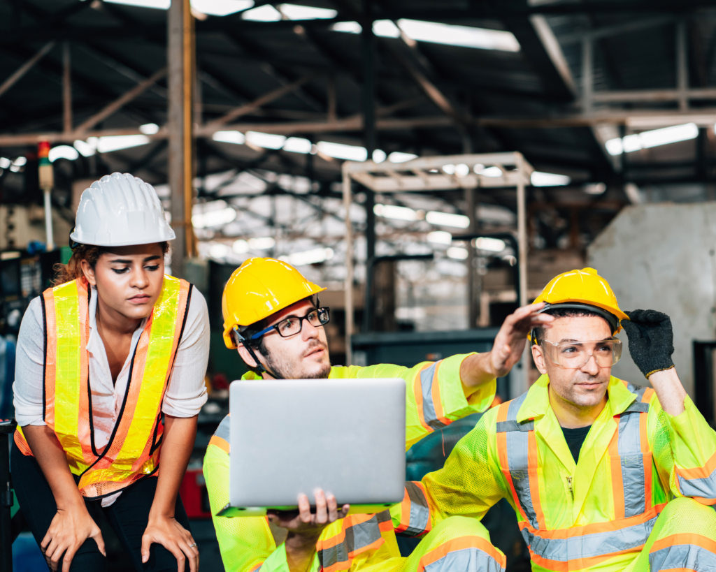 three people wearing a hi visibility vest and overalls wearing hard hats, pointing at the distance. The middle person holding at the laptop